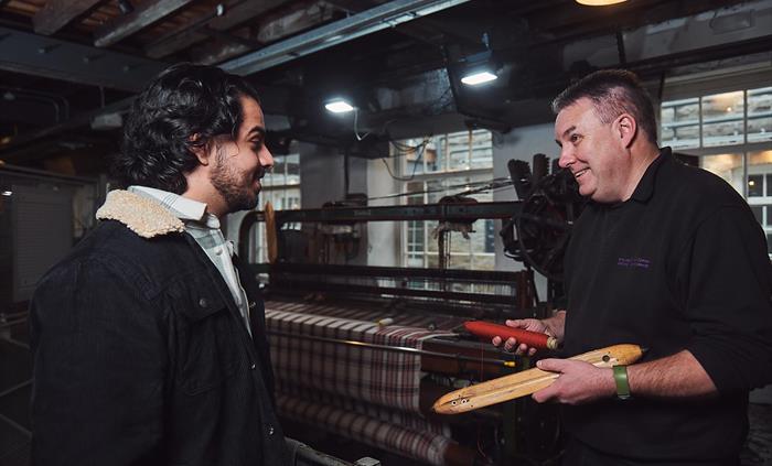 A visitor talks to a craftsman in the weaving workshop. The craftsman explains the weaving process. He is holding a bobbin of red thread and a wooden